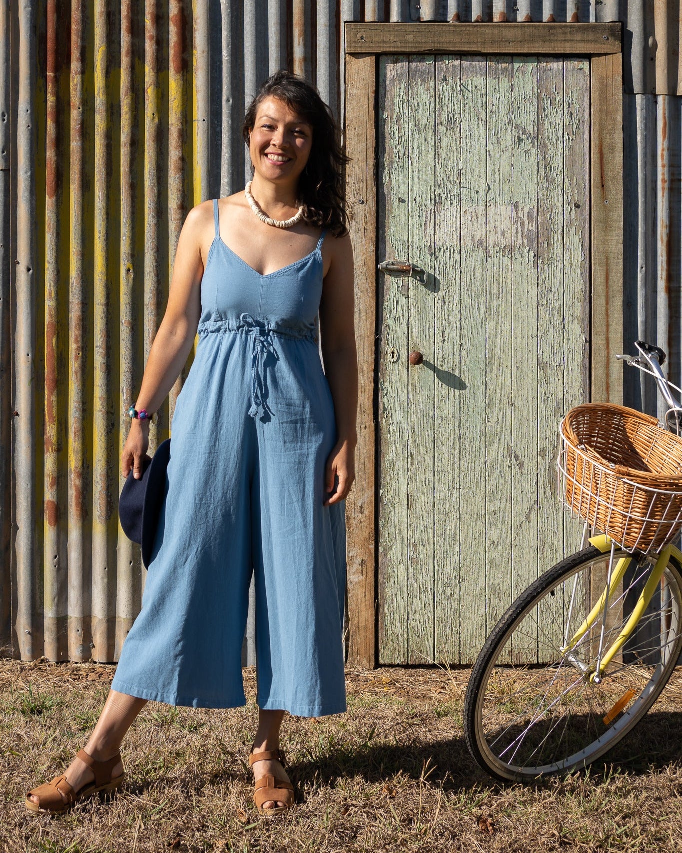 A person wearing a sky blue romper with a shirred elastic back and adjustable length straps, standing next to a yellow bicycle.