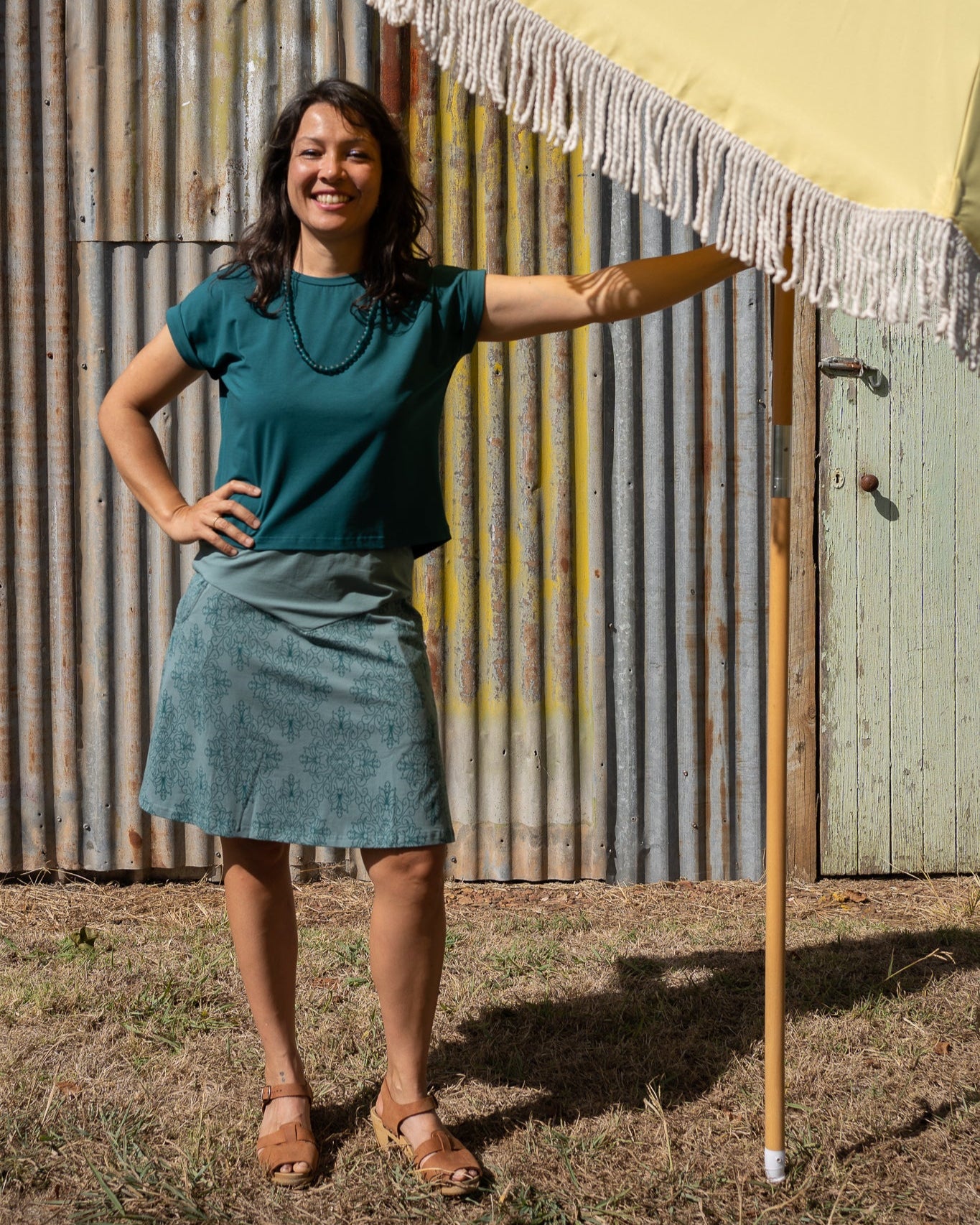 Woman standing under a yellow umbrella with a fringed edge, wearing a green top and blue skirt, against a corrugated metal wall.