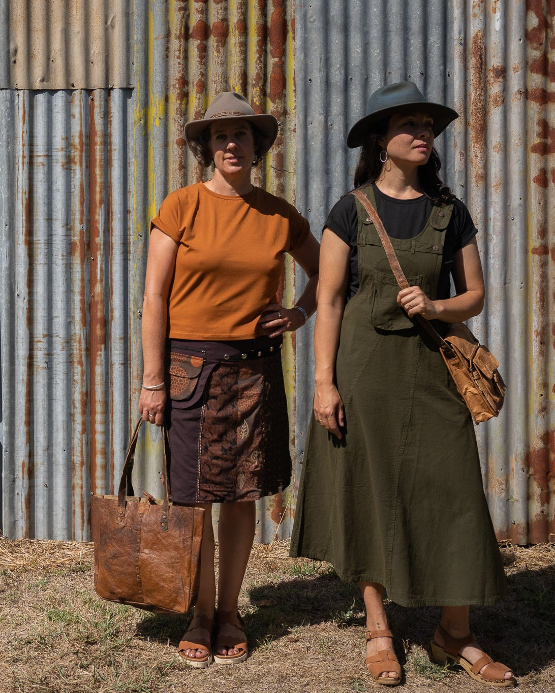 Two women standing in front of a corrugated metal building, wearing hats and carrying bags.