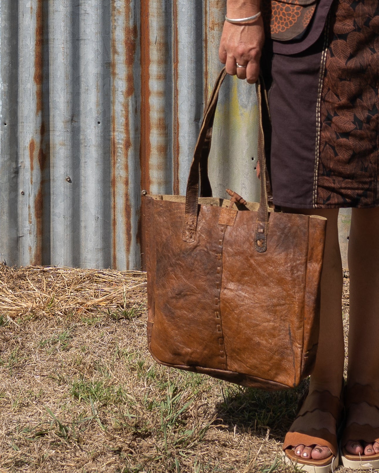 Person holding a brown leather tote bag against a rustic metal wall.