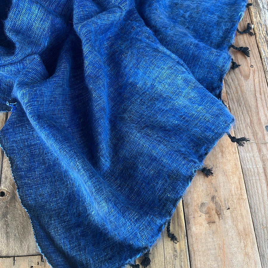 A textured royal blue woven scarf with black tassels draped over rustic wooden planks.