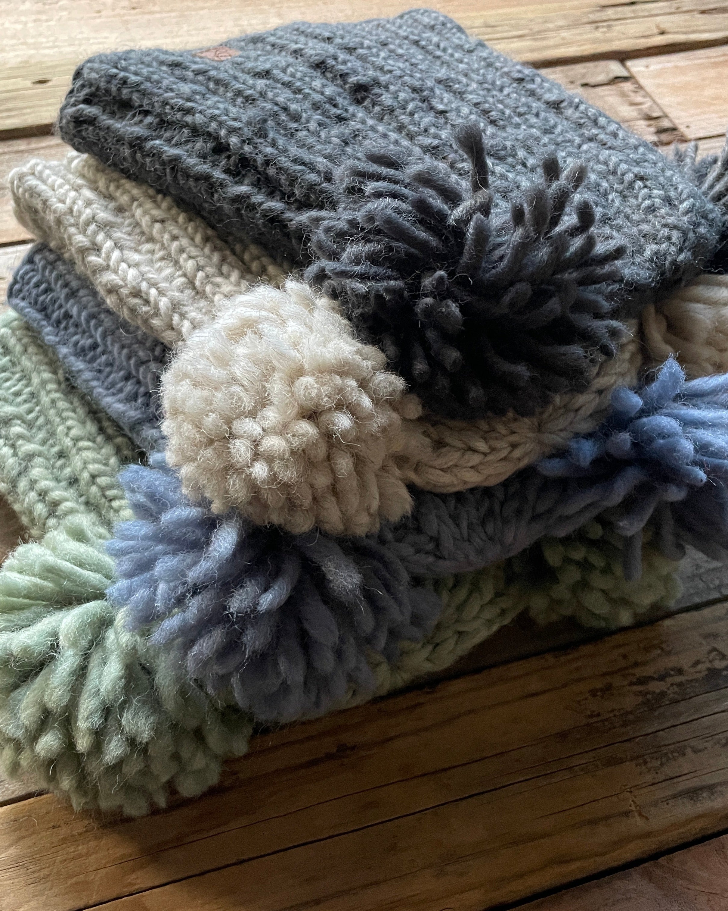 Stack of knitted hats with 2 pom-poms on a wooden surface