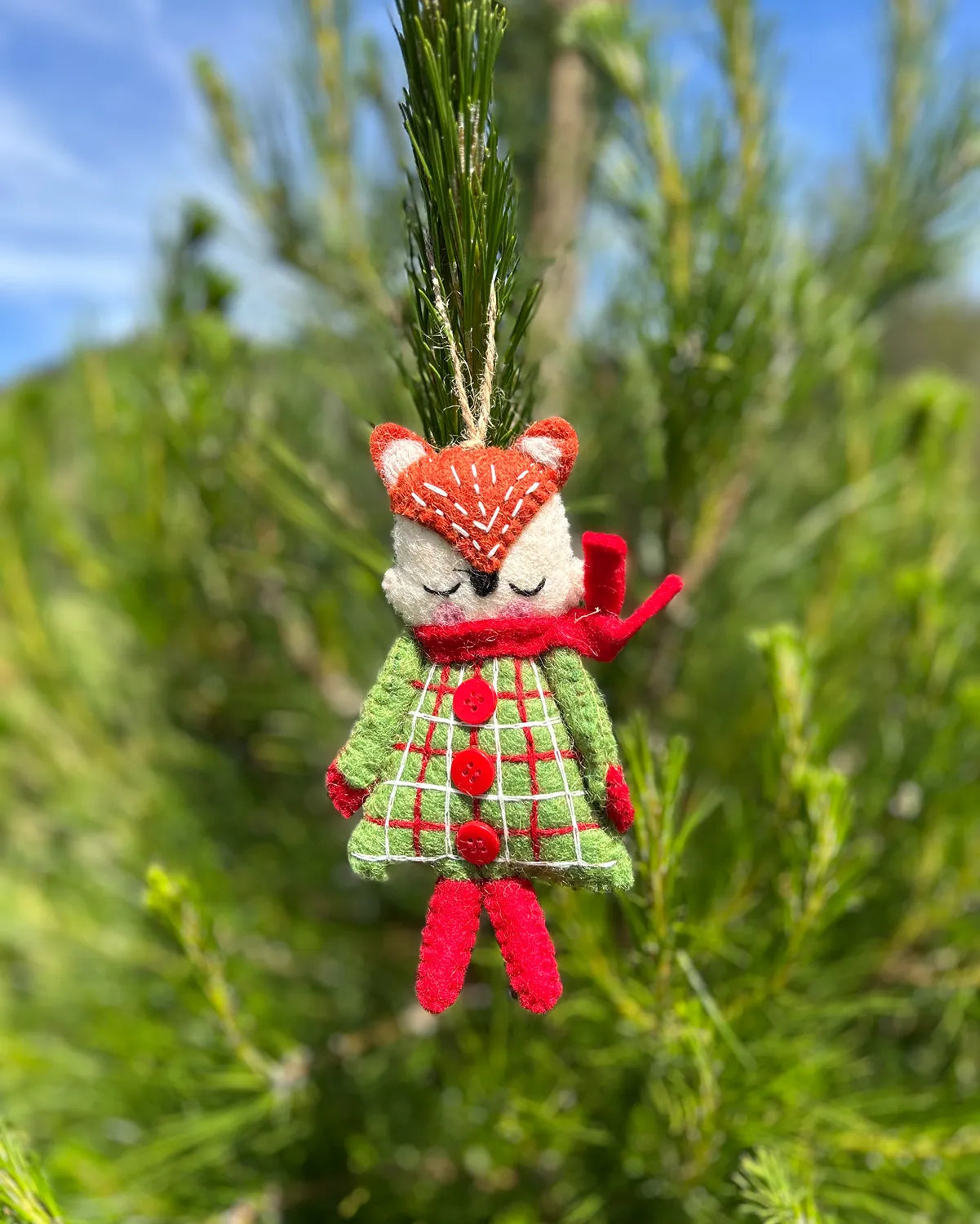 Decorative fox ornament with a green plaid dress and red accents hanging on a tree branch.