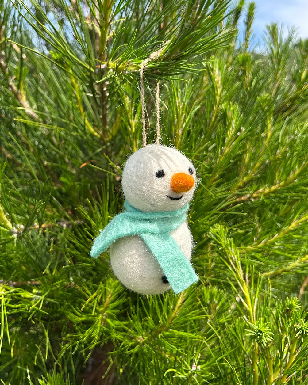 Small snowman ornament with a green scarf hanging in a pine tree.