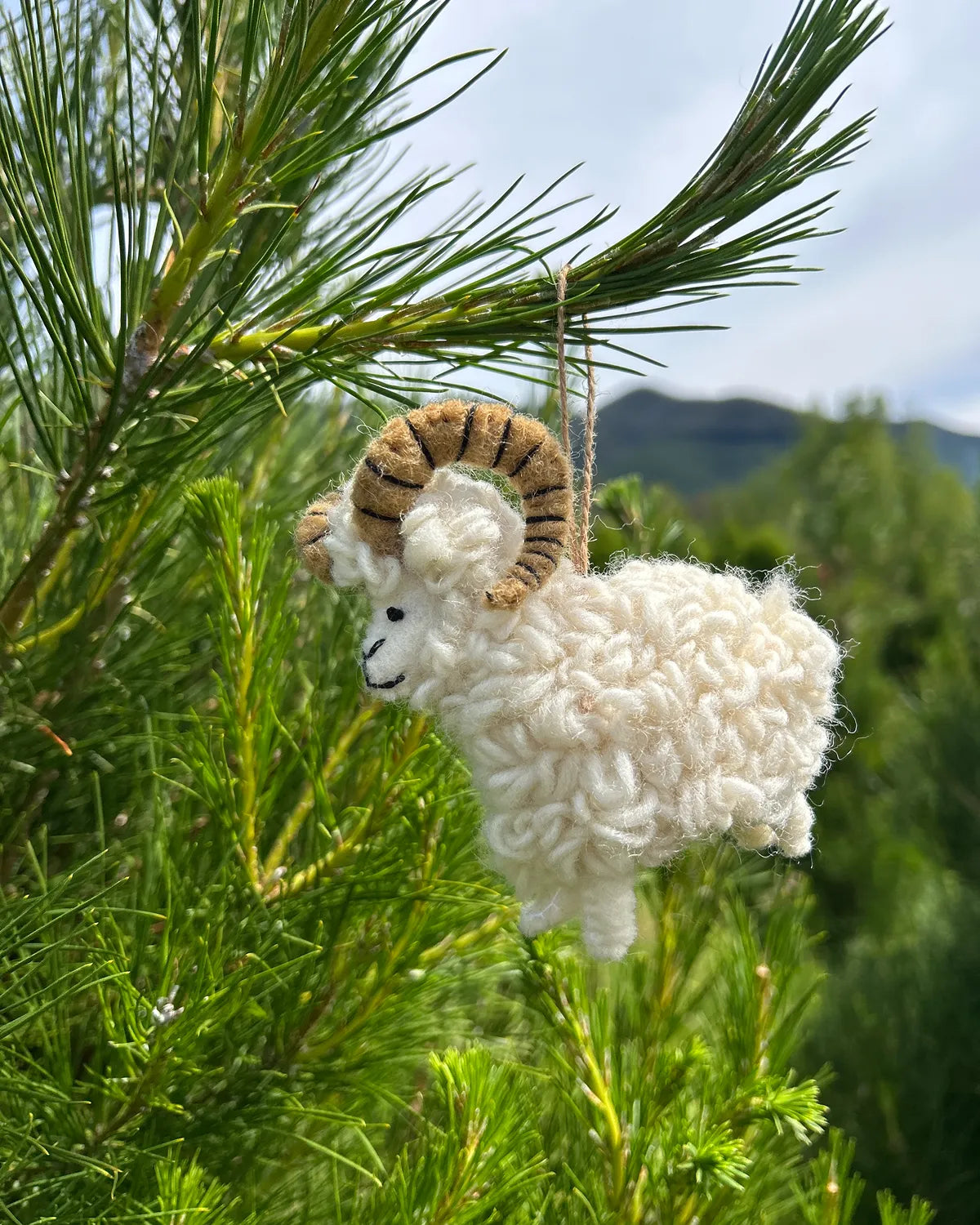 White sheep ornament hanging on a pine branch with a mountainous background