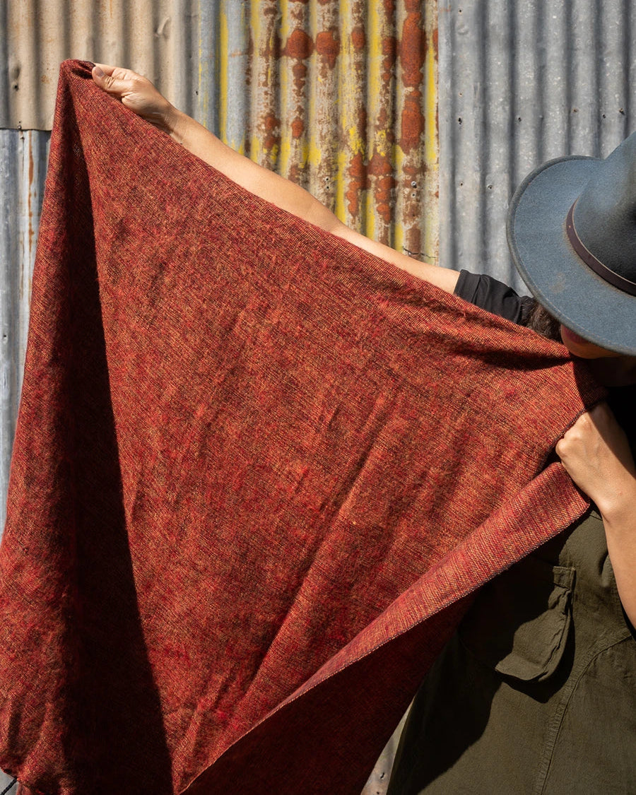 A person wearing a dark hat holds up a large rust-red woven , shawl, or scarf, against a corrugated metal wall with rust patches.