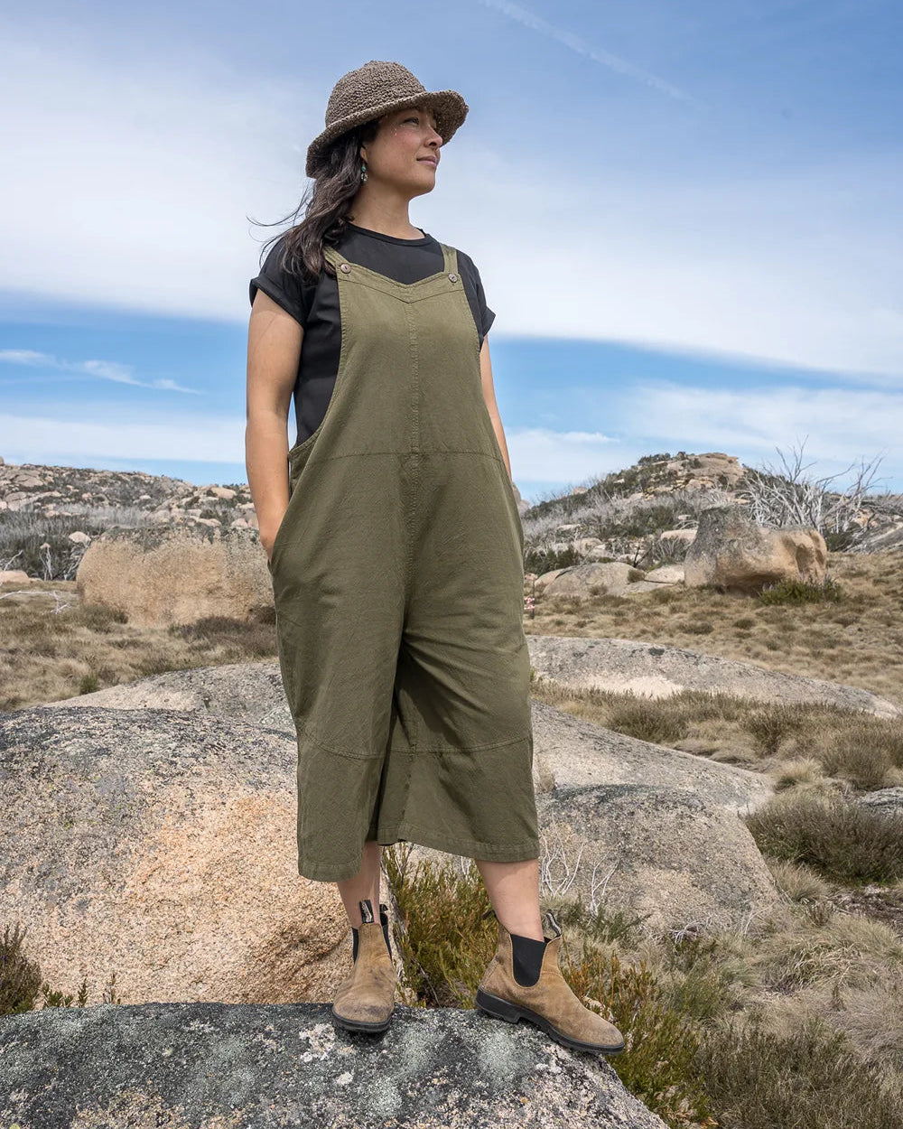 Woman in a green jumpsuit and hat standing on a rocky landscape with a blue sky.
