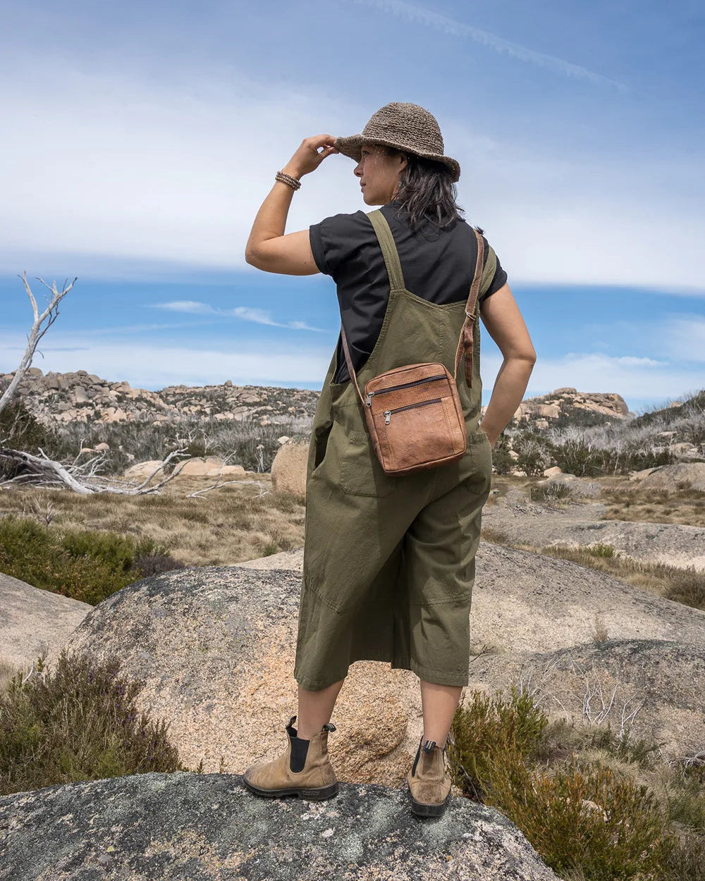 Person in green overalls and hat standing on a rocky landscape with a blue sky.
