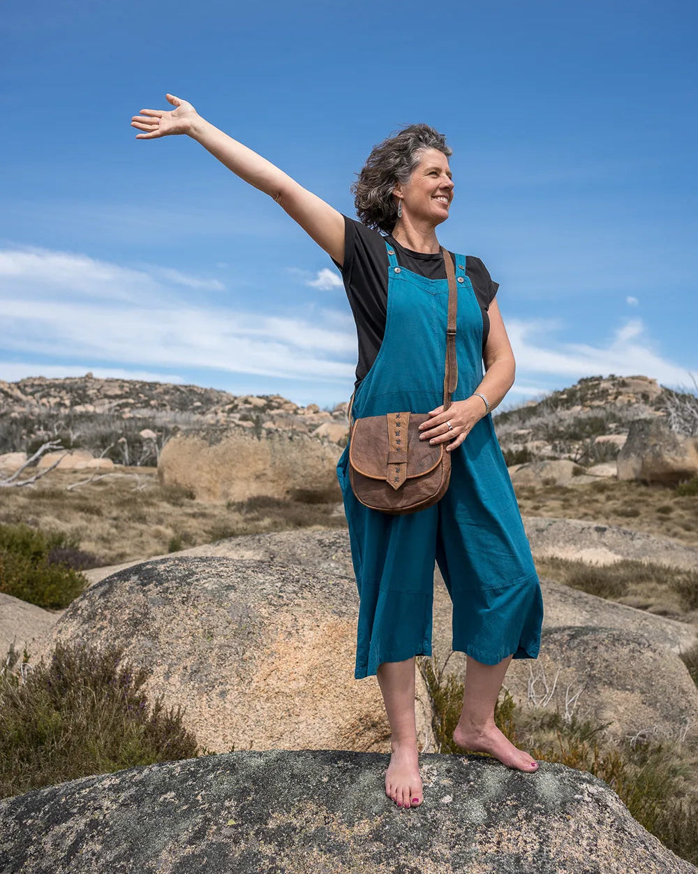 Woman in a blue overalls on a rock with arms outstretched against a blue sky.