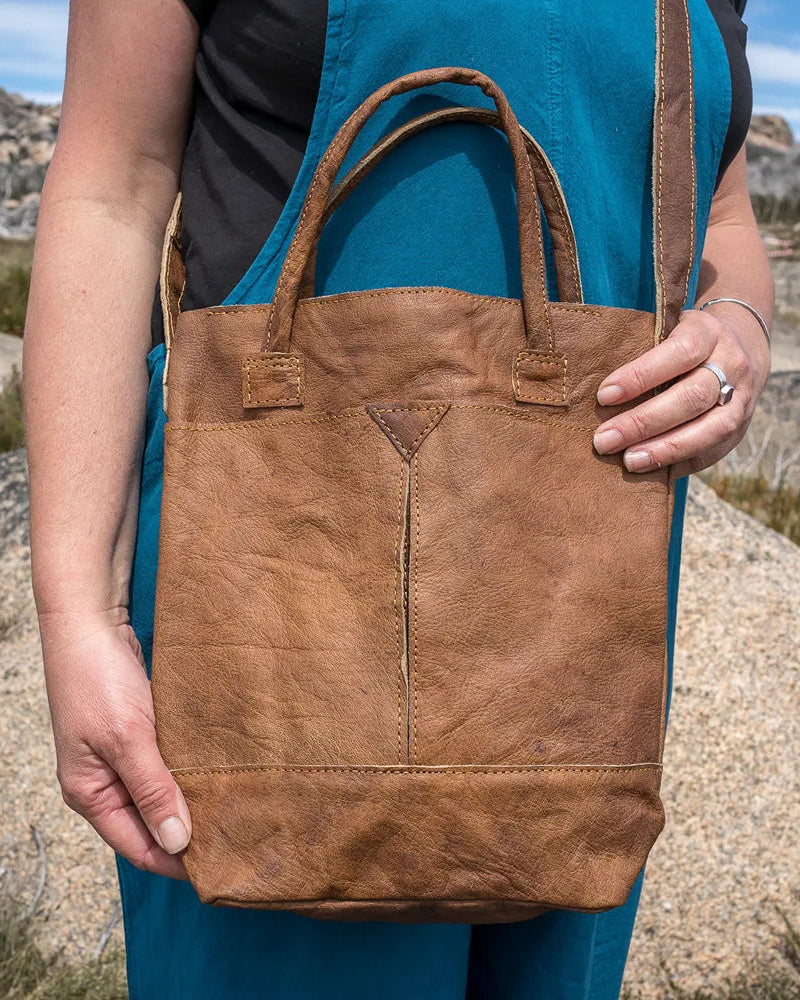 Person holding a brown leather tote bag in a desert-like landscape