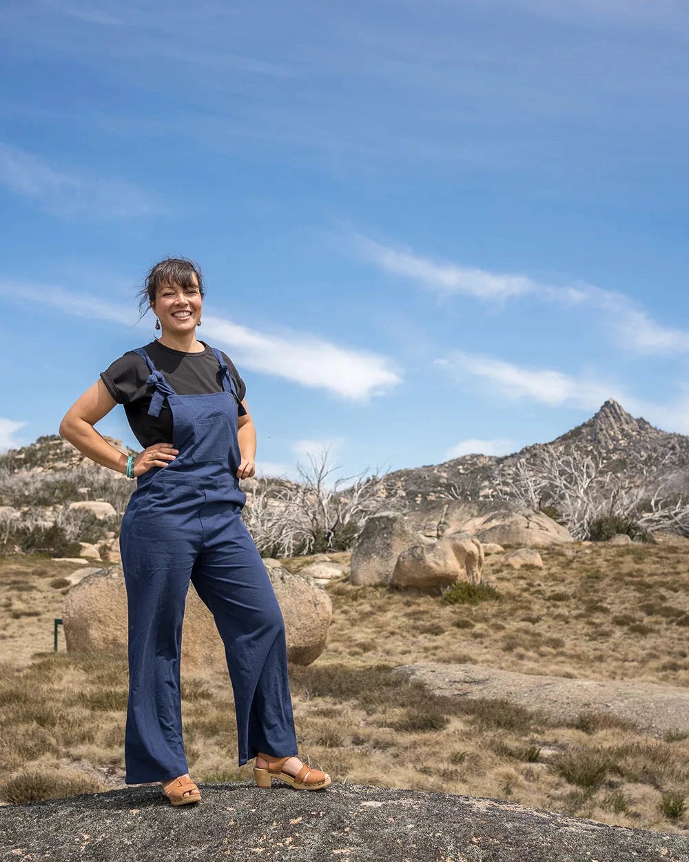Person wearing blue overalls standing on a rocky outcrop with mountains in the background