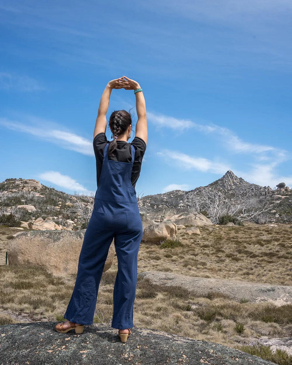 Person in blue overalls standing on a rock with mountains and clear sky in the background