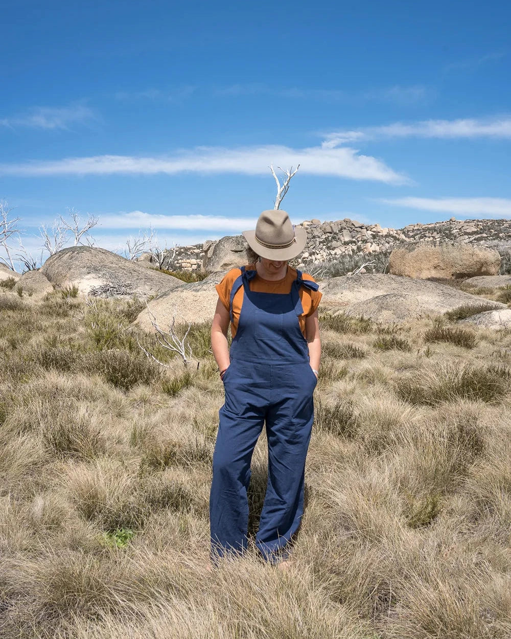 Person wearing a hat and blue overalls standing in a rocky, grassy landscape with a clear blue sky.