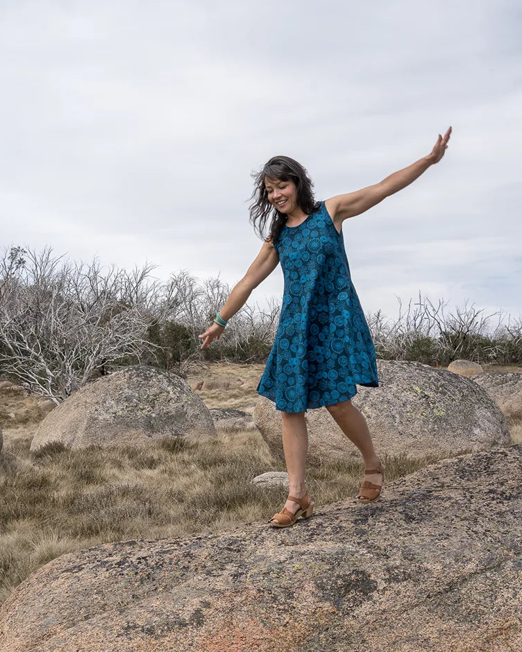 Woman in a blue dress standing on rocks with arms outstretched in an open landscape