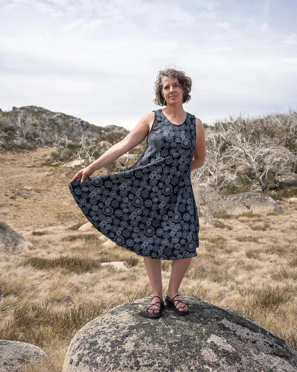 Woman in a sleeveless black print dress standing on a rock in a natural landscape