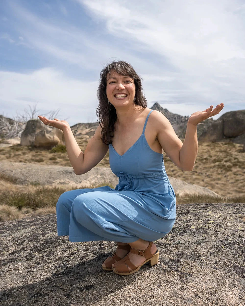 Woman in a blue dress squatting outdoors with a clear sky and rocky landscape.