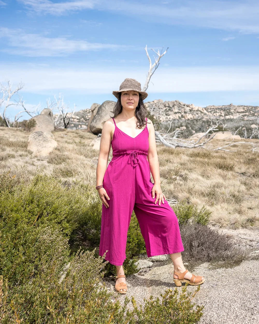 Woman in a pink jumpsuit and hat standing in a desert landscape with blue sky.