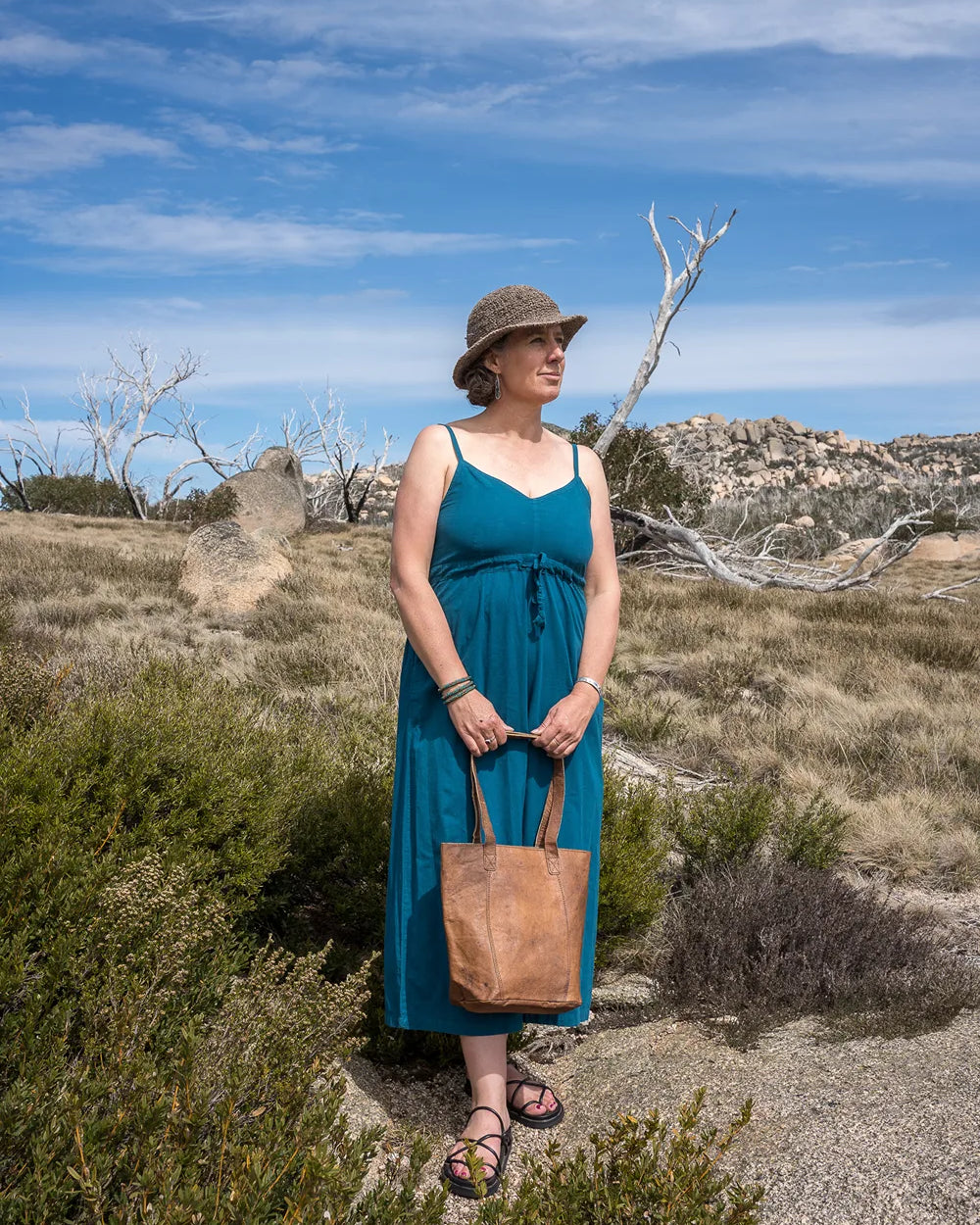 Woman in a blue onepiece and hat standing in a desert landscape with a brown bag.