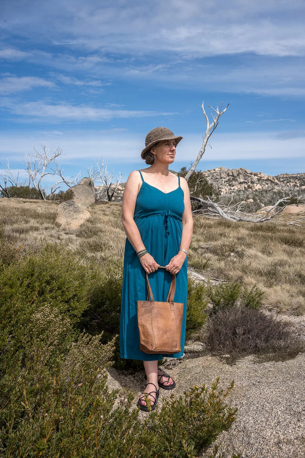 Woman in a blue onepiece and hat standing in a desert landscape with a brown bag.