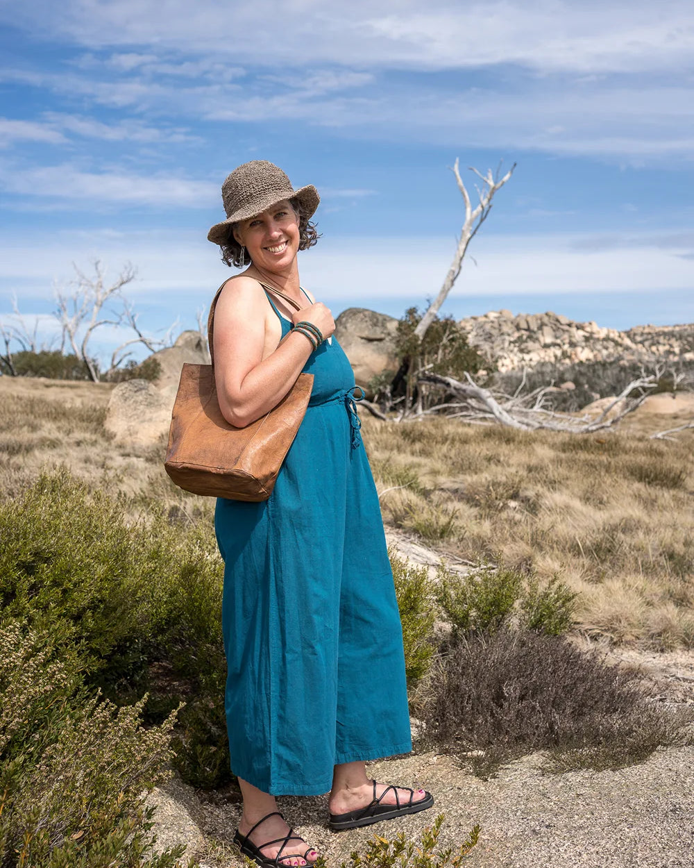 Woman in a blue dress and hat standing in a desert landscape with a brown bag.