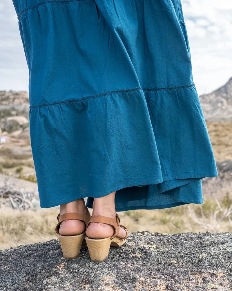 Person wearing a blue tiered skirt and brown shoes standing on a rocky landscape with mountains in the background.