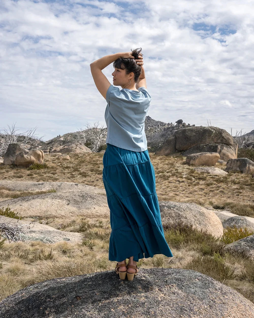Person standing in a blue ruffle skirt with a scenic background of mountains and sky.