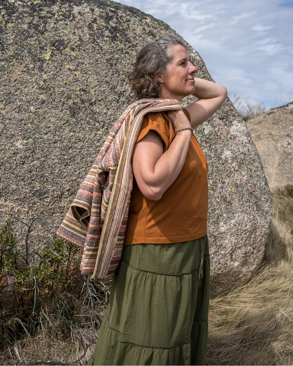 Woman in rust and khaki colour clothes standing outdoors with a large rock and blue sky in the background