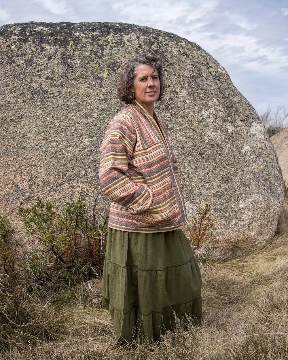 Person wearing a patterned jacket and green skirt standing in front of a large rock with a blue sky.