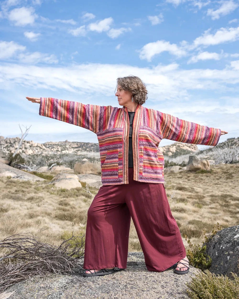Person wearing a colorful jacket and maroon pants standing in a rocky landscape with blue sky.