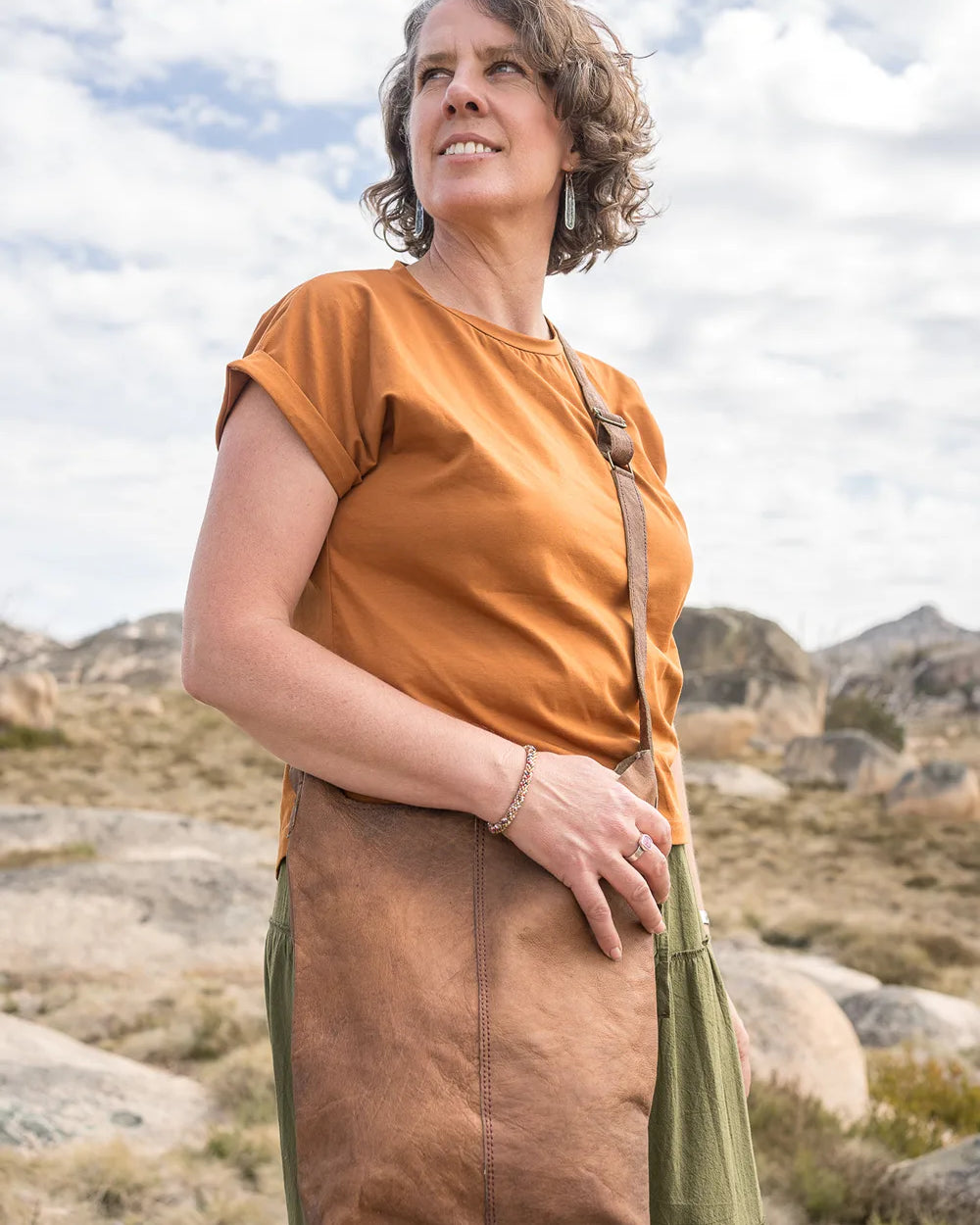 Woman in an orange shirt and brown apron standing in a rocky landscape with mountains in the background.