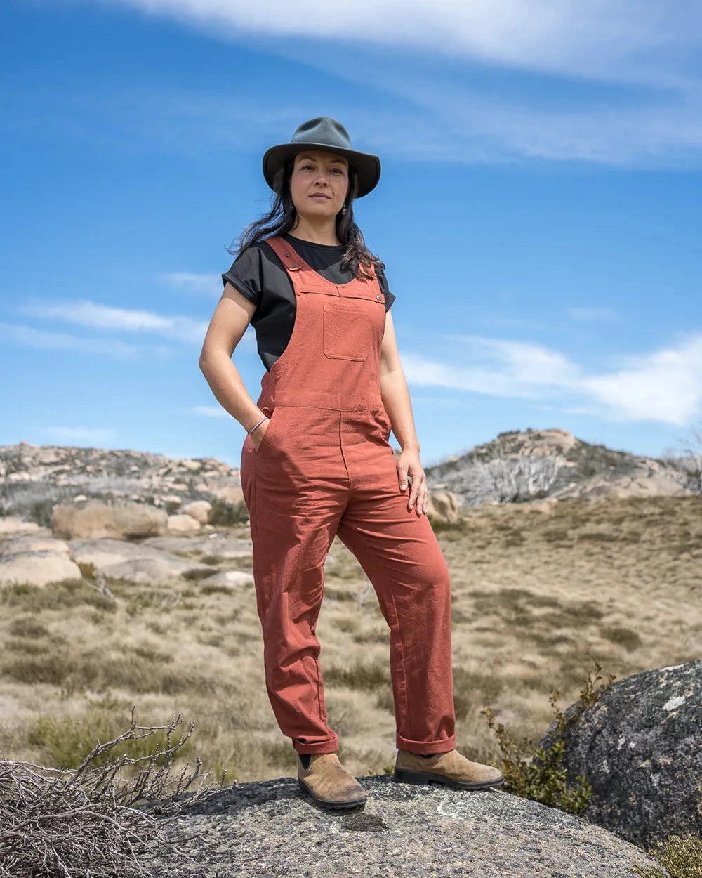 A woman stands outdoors on a rocky hillside wearing rust-colored bib overalls over a black t-shirt, a dark wide-brim hat, and brown boots, with a dry grassy landscape and blue sky in the background.