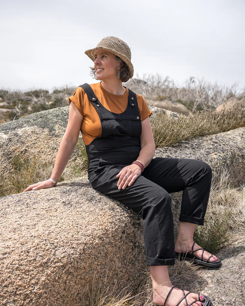 A woman wearing a straw hat, a mustard yellow short-sleeve top, and black overall-style pants sits on a large rock in an outdoor rocky landscape under a bright sky.