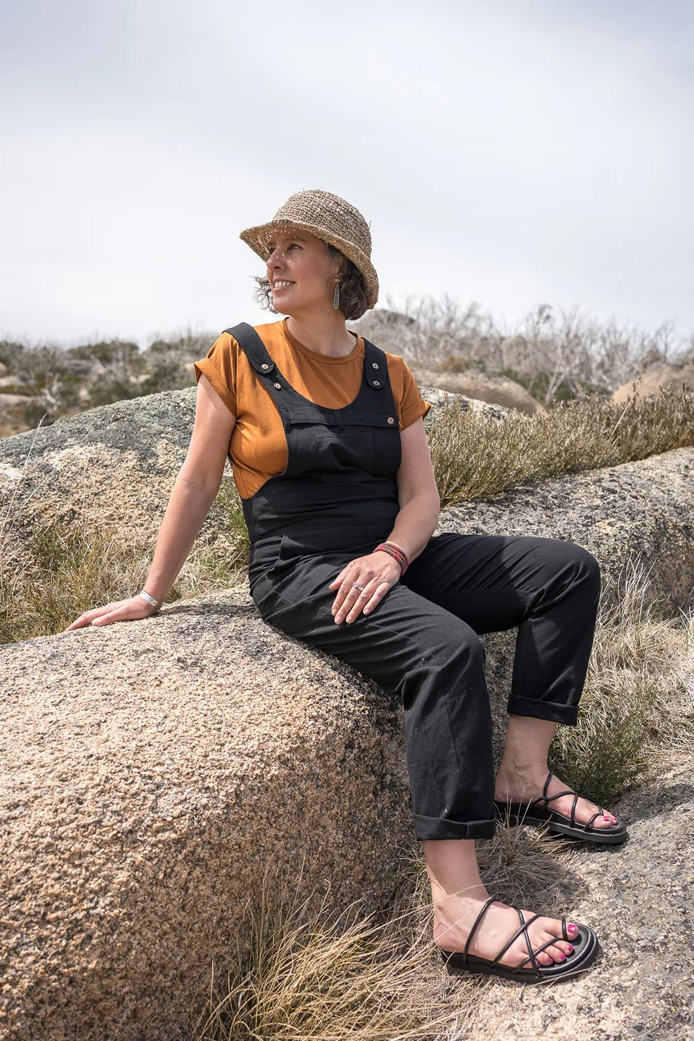 A woman wearing a straw hat, a mustard yellow short-sleeve top, and black overall-style pants sits on a large rock in an outdoor rocky landscape under a bright sky.