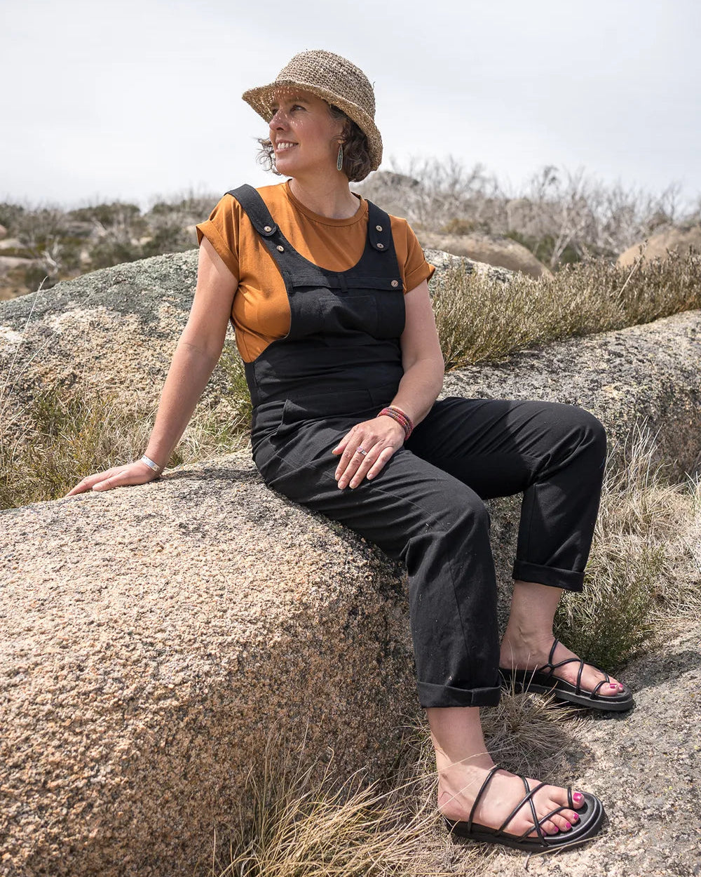 A woman wearing a straw hat, a mustard yellow short-sleeve top, and black overall-style pants sits on a large rock in an outdoor rocky landscape under a bright sky.