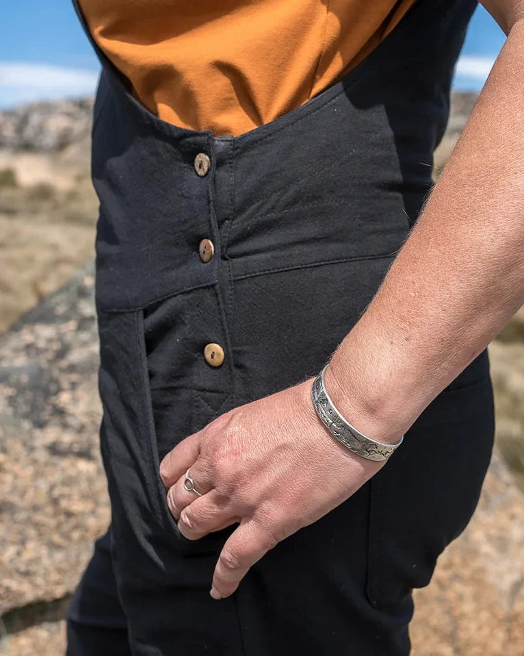 A close-up lifestyle shot of a person wearing black bib overalls over a mustard orange sleeveless top, standing outdoors in a rocky desert landscape under a blue sky.
