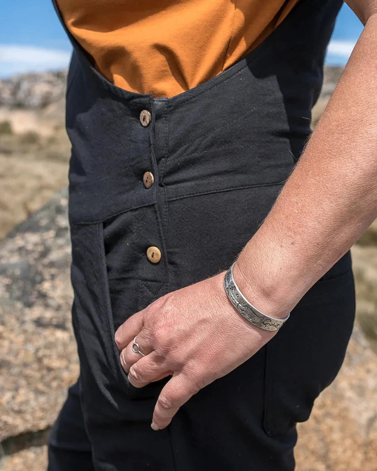A close-up lifestyle shot of a person wearing black bib overalls over a mustard orange sleeveless top, standing outdoors in a rocky desert landscape under a blue sky.
