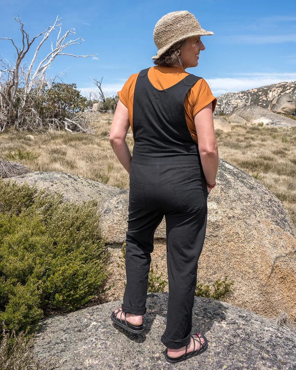 A woman standing outdoors on a rocky hillside, viewed from behind, wearing a black sleeveless jumpsuit or romper over an orange short-sleeve top, with a straw hat and sandals.