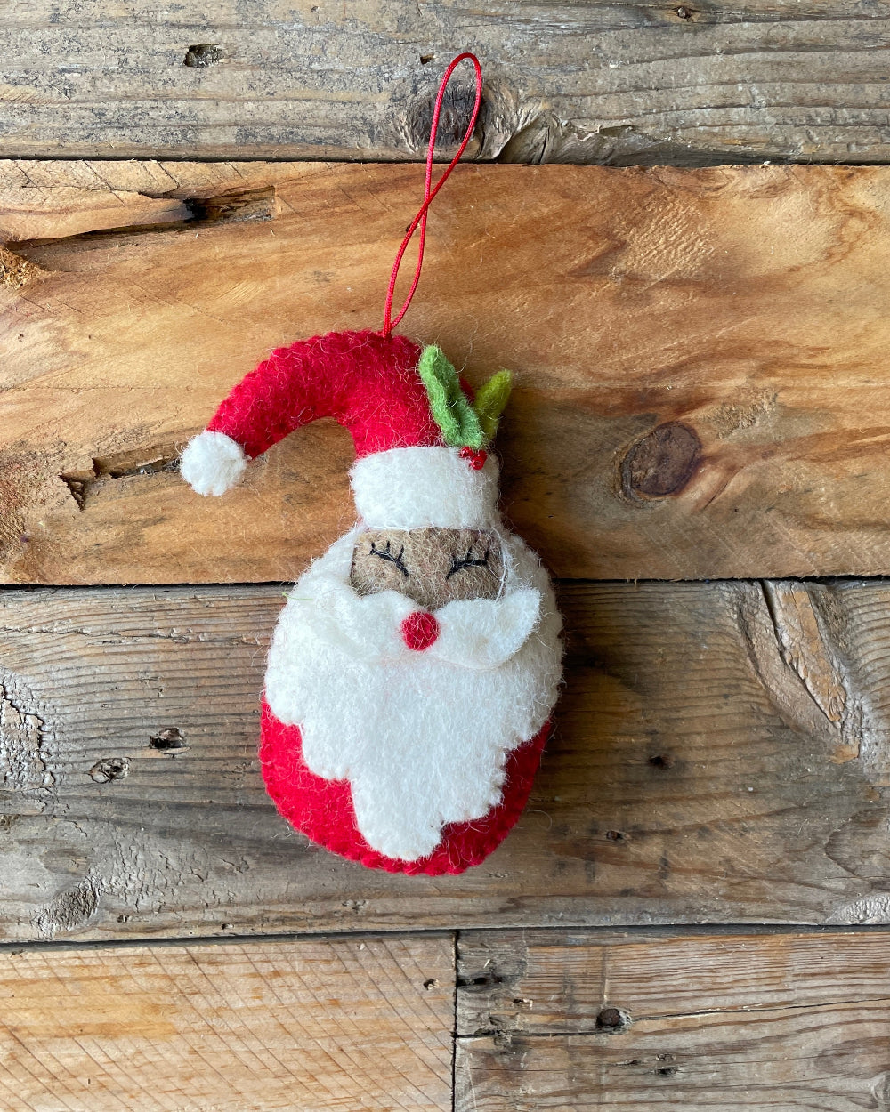 A red and white felt Santa Claus Christmas tree decoration hanging against a wooden background.