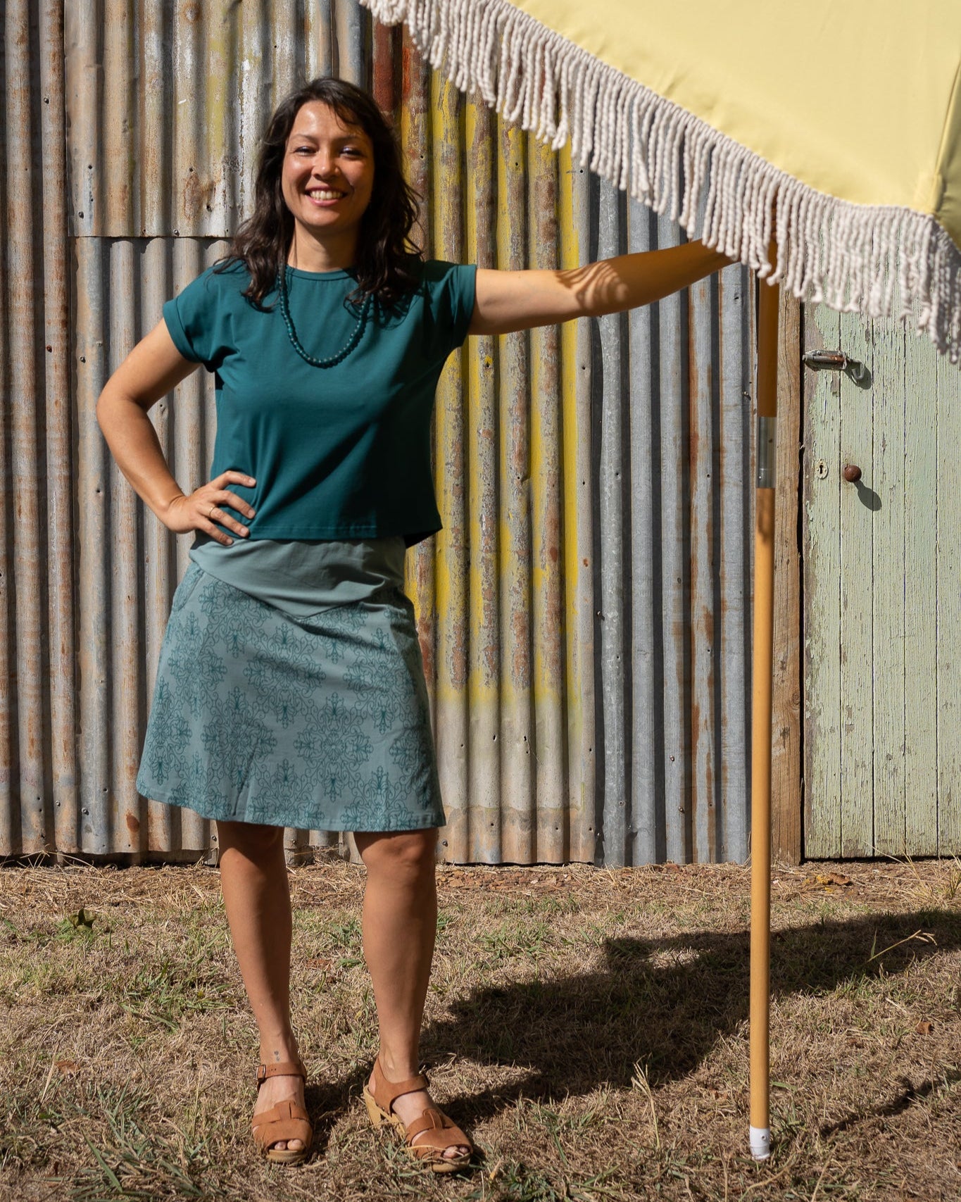 Woman standing under a yellow umbrella with a fringed edge, wearing a green top and blue skirt, against a corrugated metal wall.