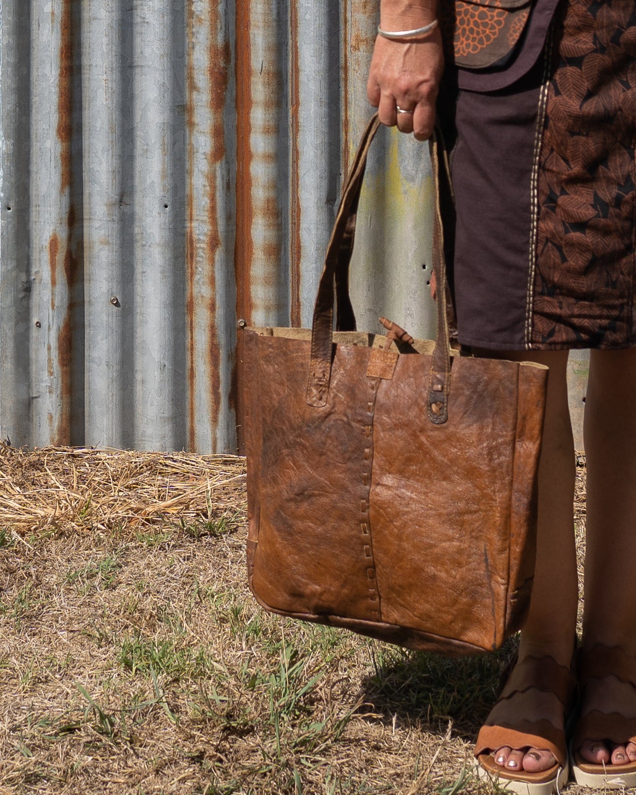 Person holding a brown leather tote bag against a rustic metal wall.
