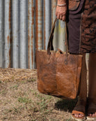 Person holding a brown leather tote bag against a rustic metal wall.