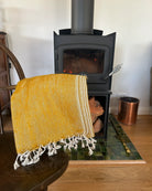 A folded mustard yellow woven throw blanket with white fringe is draped over a dark wooden chair in front of a black wood-burning stove. The scene is set in a home interior with a tiled hearth and wood floor.