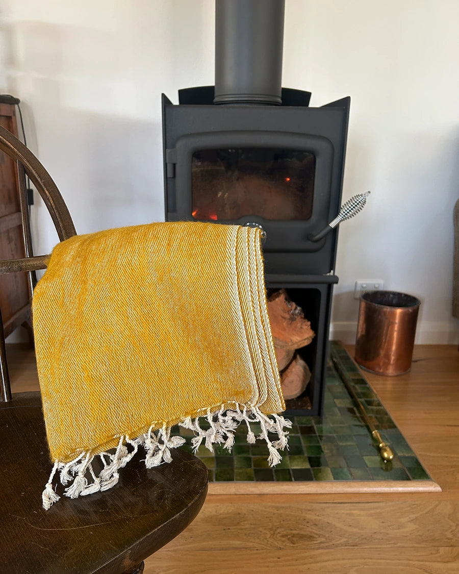 A folded mustard yellow woven throw blanket with white fringe is draped over a dark wooden chair in front of a black wood-burning stove. The scene is set in a home interior with a tiled hearth and wood floor.