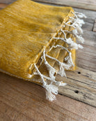 A close-up of a mustard yellow woven textile with white tassels, laid diagonally on a rustic wooden surface.