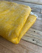 A folded mustard yellow woven textile, likely a scarf or throw blanket, displayed on a rustic wooden surface.