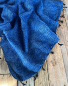 A textured royal blue woven scarf with black tassels draped over rustic wooden planks.