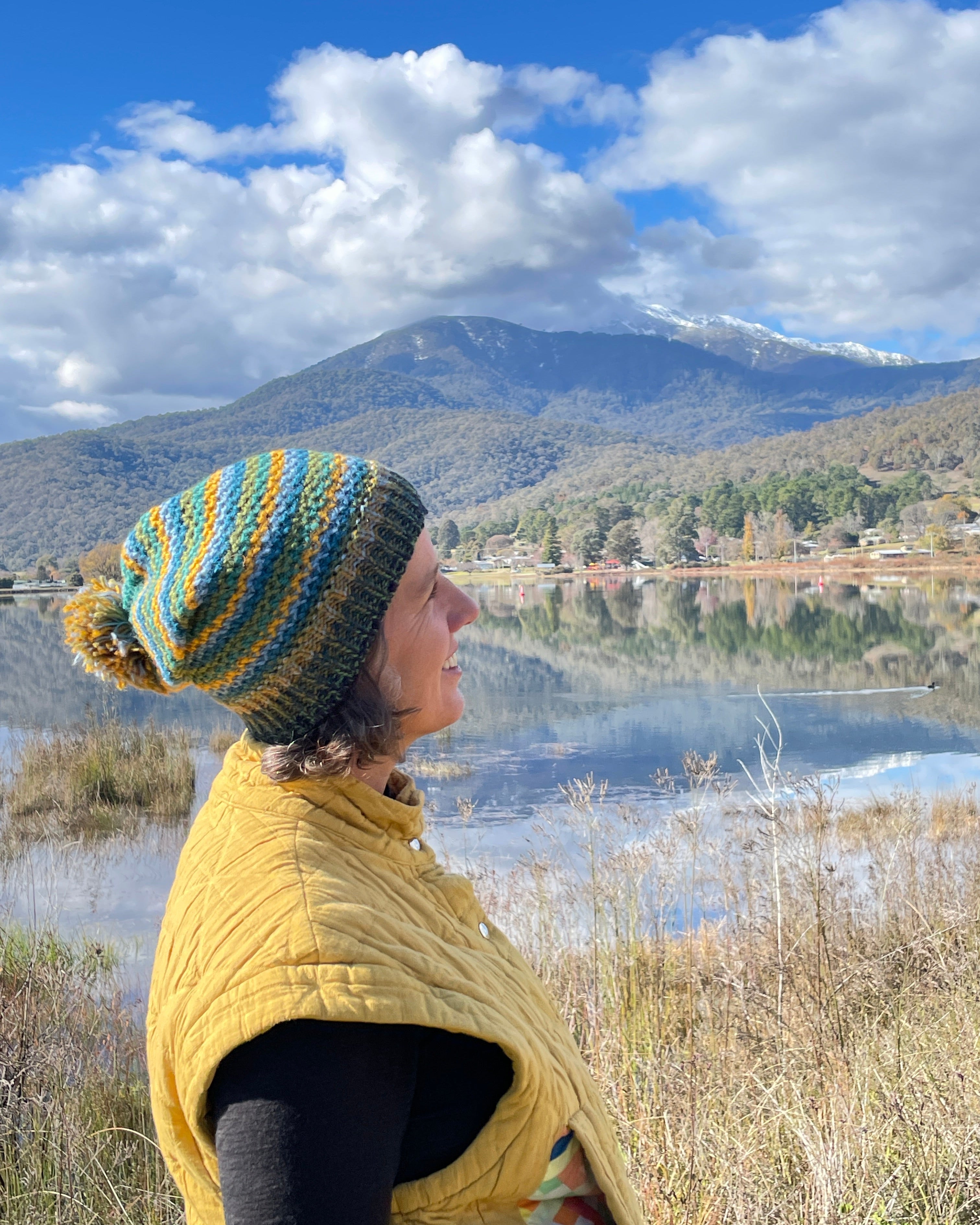 Person wearing a colorful knitted hat and yellow jacket standing in a field with mountains in the background.