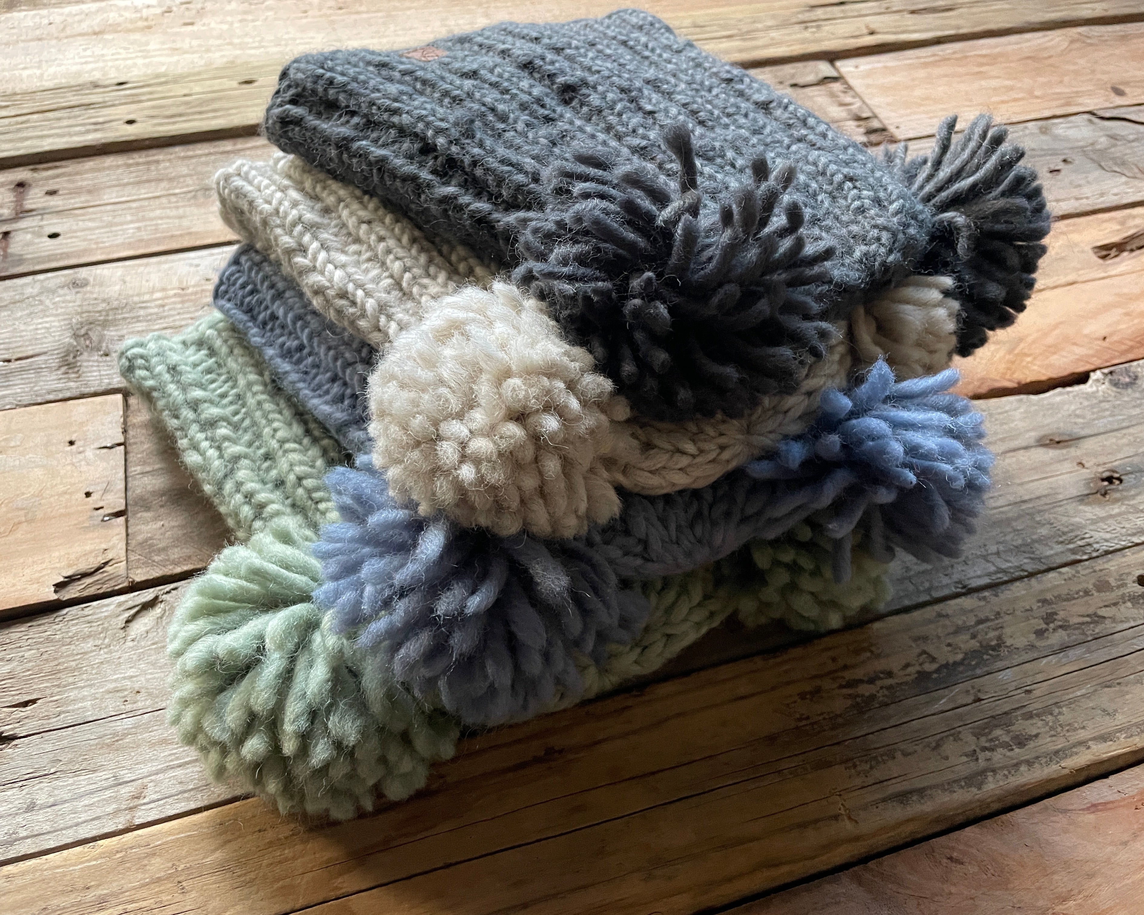 Stack of knitted hats with 2 pom-poms on a wooden surface