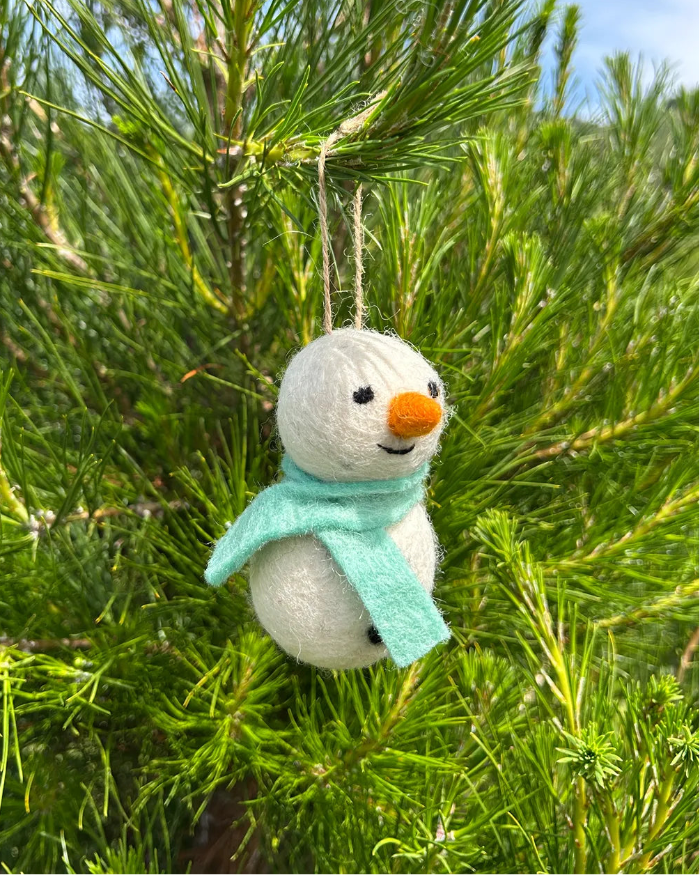 Small snowman ornament with a green scarf hanging in a pine tree.
