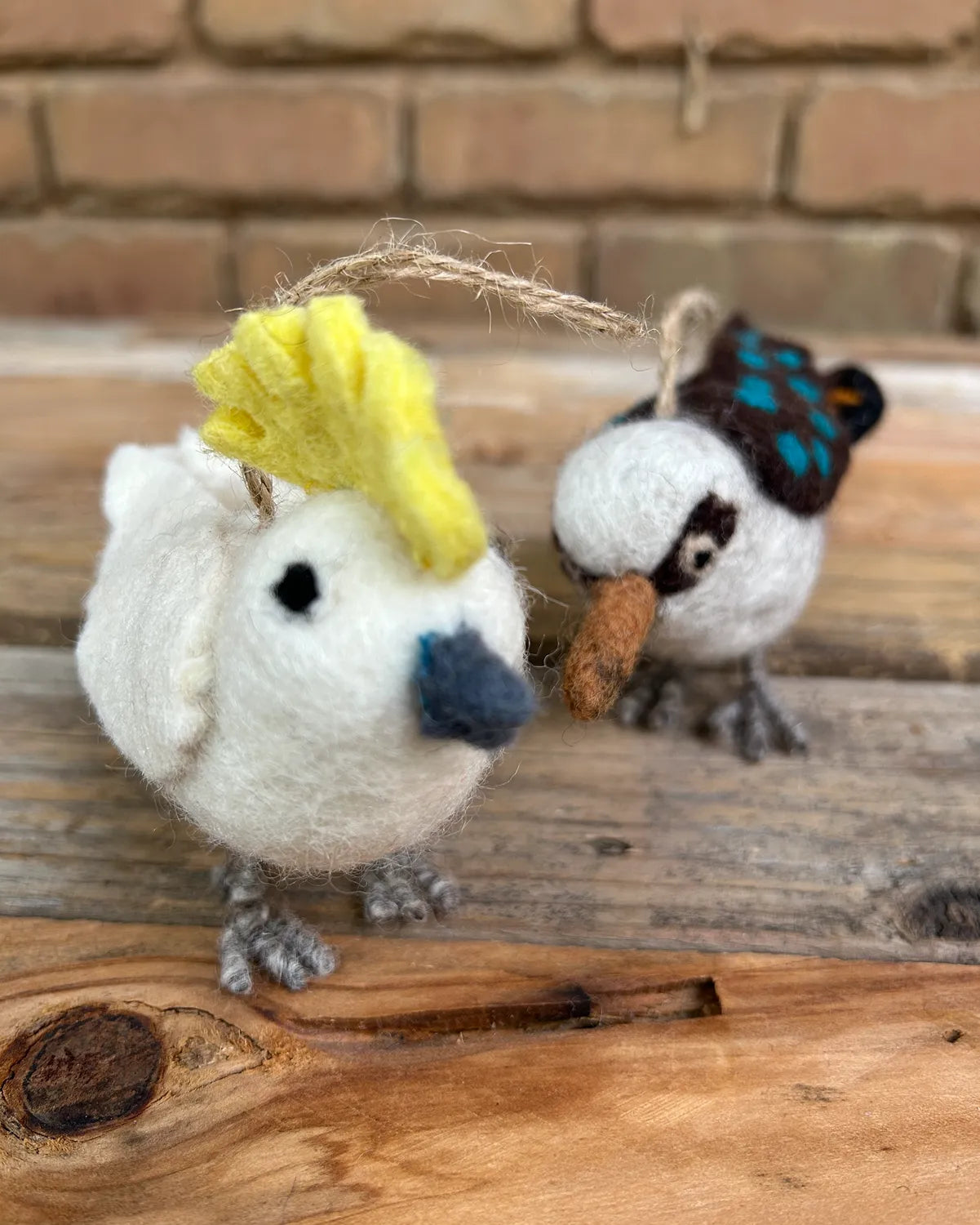 Two needle-felted birds on a wooden surface with a brick wall background